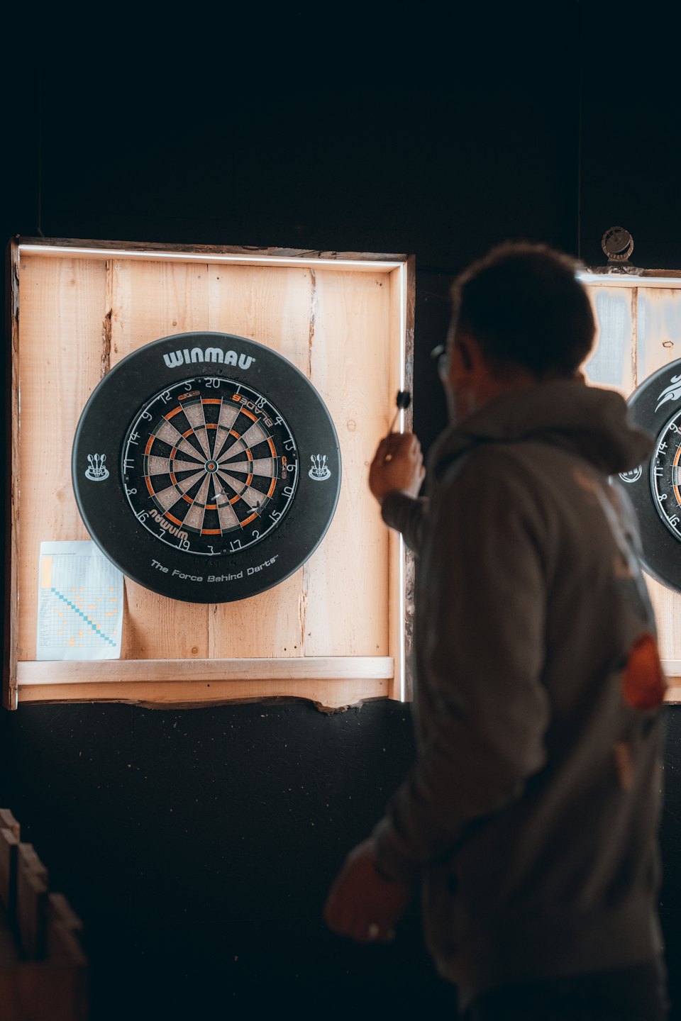 Eine Person zielt mit einem Dartpfeil auf eine Dartscheibe an einer Holzwand.